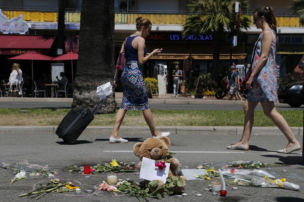 A woman pulls her suitcase as she walks past flowers and a stuffed toy placed in tribute to victims of the truck attack along the Promenade des Anglais on Bastille Day that killed scores and injured as many in Nice