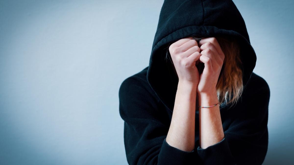 Young girl hiding her face under hooded sweatshirt isolated on b