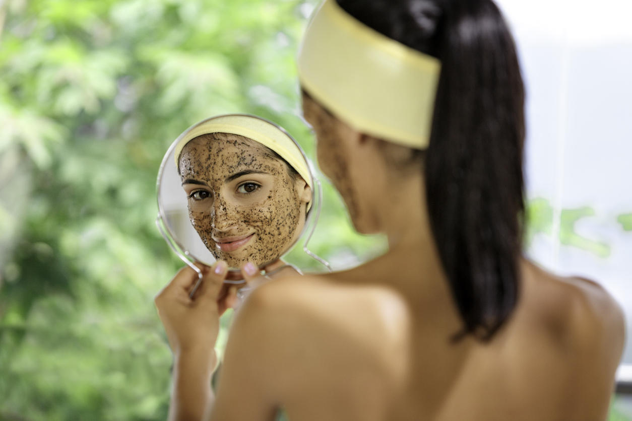 Hispanic young woman with coffe face scrub after bath looking at herself on hand mirror. Skin Exfoliation.