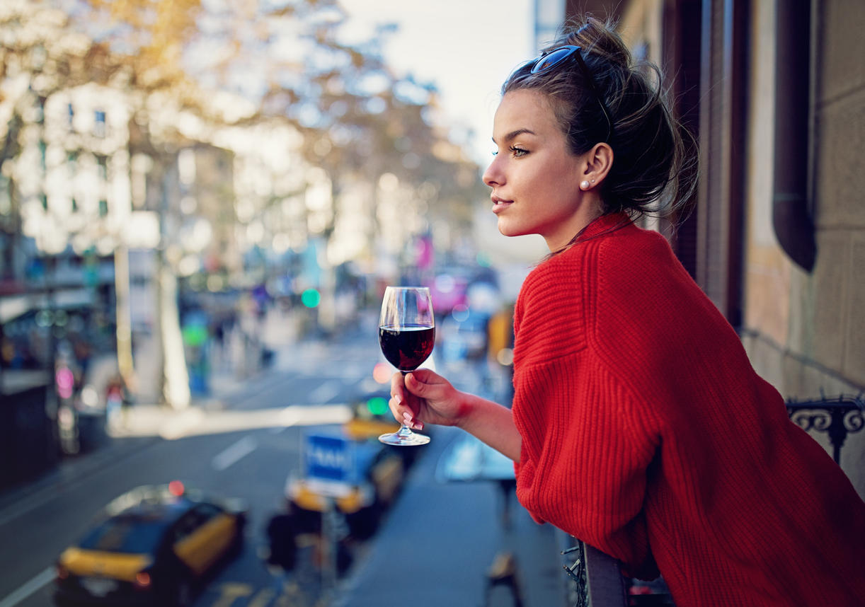 Young girl is looking the city at the balcony and drinking red wine