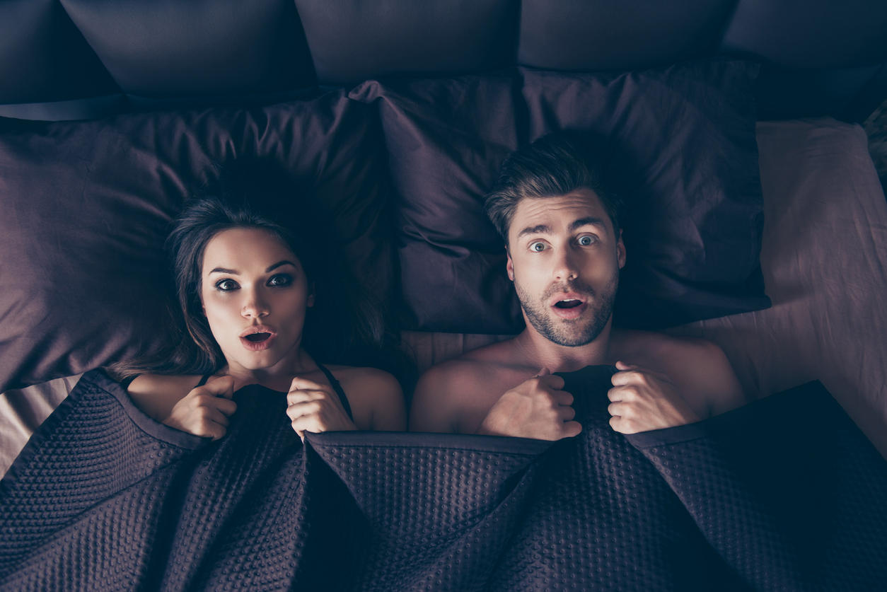 Funny lovers man and woman lying in bed covered by sheets with wide opened eyes and mouth looking at camera, having shocked and scared expressions, afraid to be found in bed by someone who unexpectedly came home from work