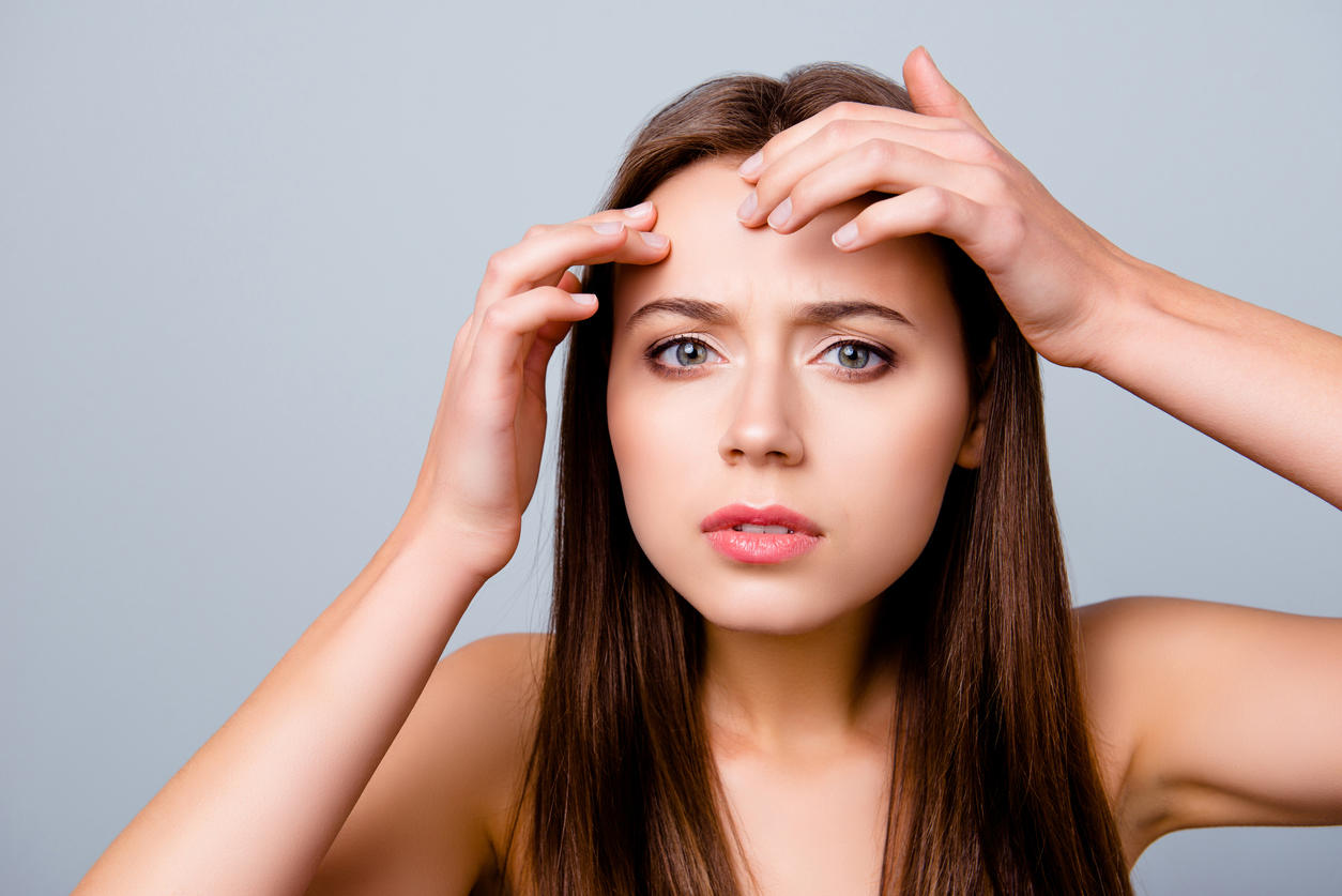 Close up portrait of frustrated sad upset beautiful young woman is squeezing out pimples on her forehead, isolated on grey background