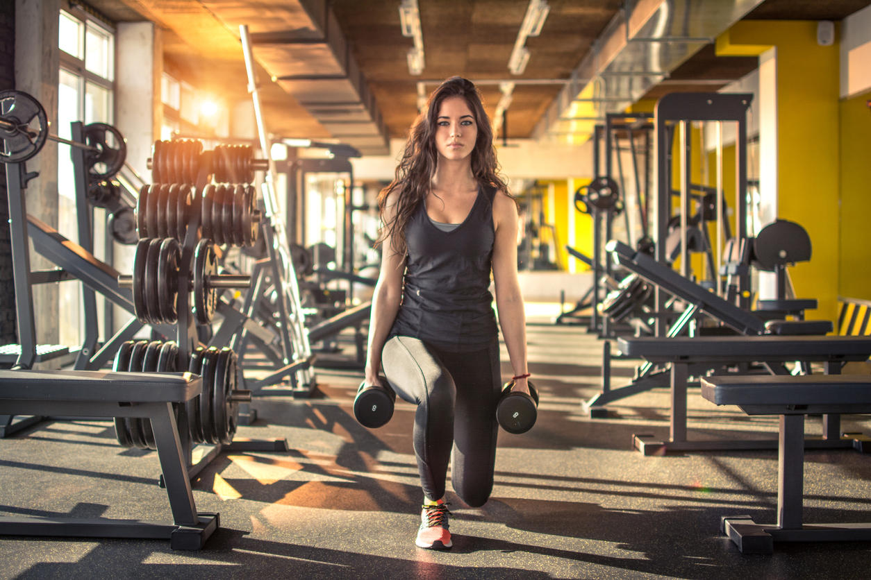 Full length portrait of young slim woman doing lunges with dumbbells in the gym.