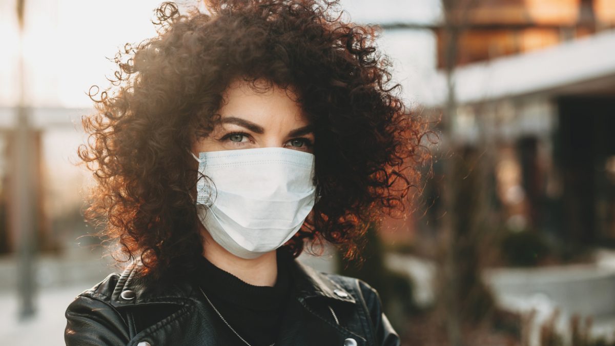 Isolated through a mask caucasian girl with nice curly hair is posing sadly outside while looking to camera