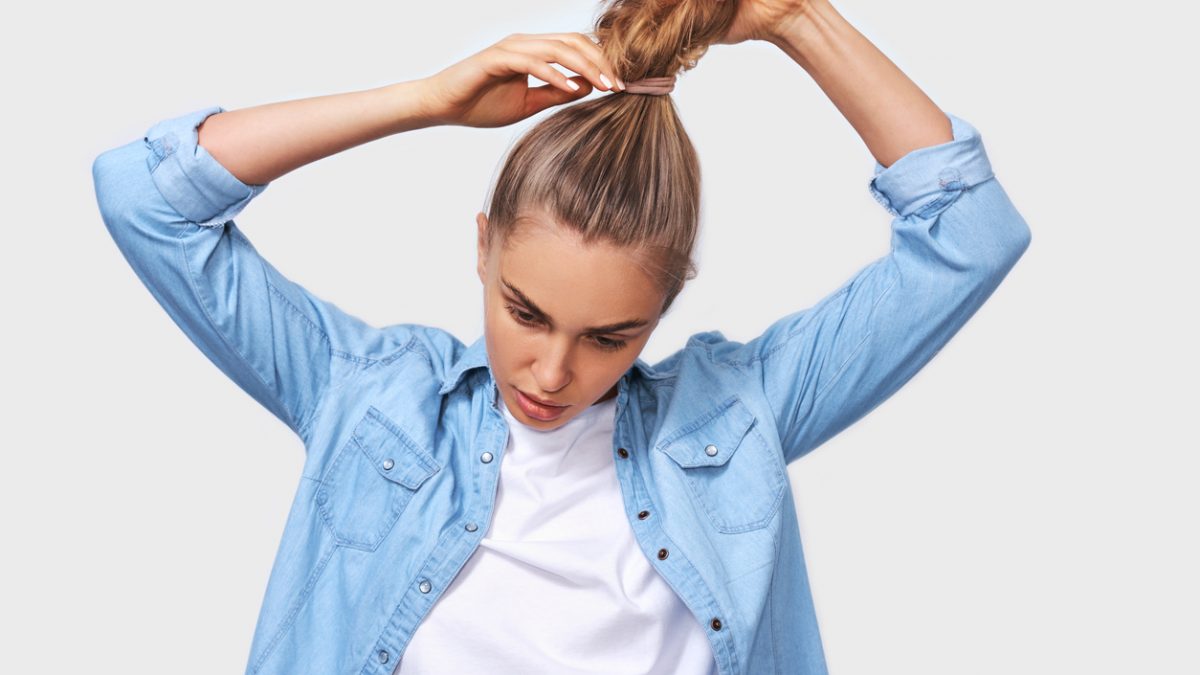 Indoor portrait of young woman collecting hair in a ponytail, wearing blue denim shirt and white t-shirt, posing over white wall. Adorable blonde female makes ponytail, advertises healthy natural hair