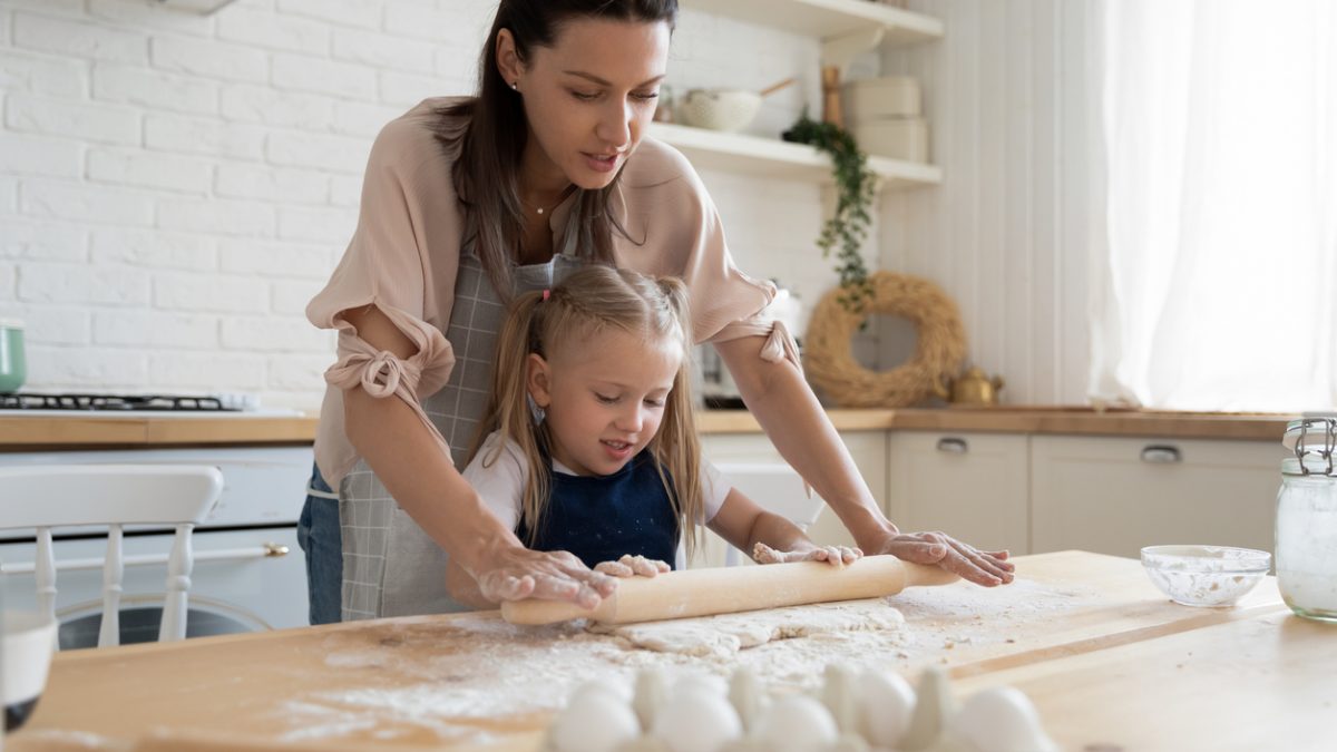 Young mother showing preschool daughter how to roll out dough.