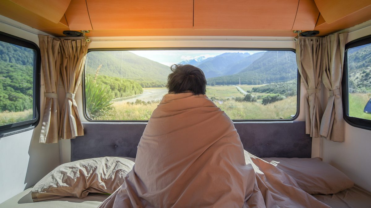 Young Asian man traveler staying in the blanket looking at mountain scenery through the window in camper van in the morning. Road trip in summer of South Island, New Zealand.