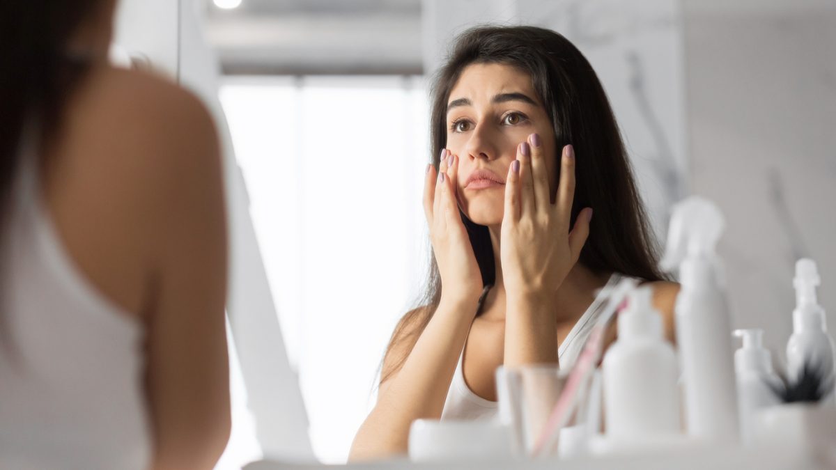 Woman Touching Face Looking At Skin In Mirror At Bathroom